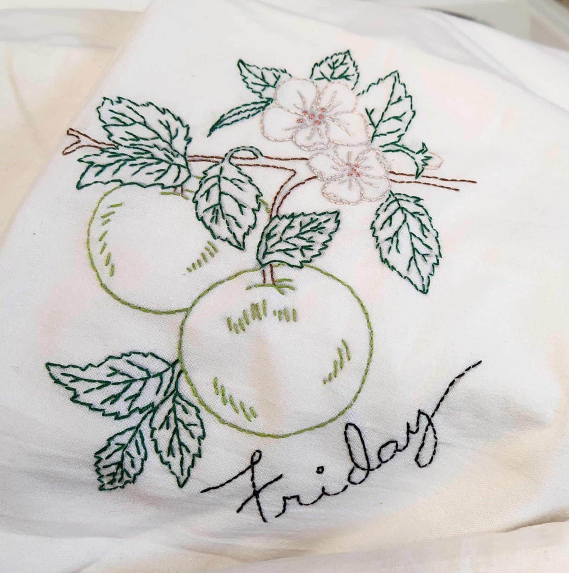 Close up on Tea Towel with an embordered Green apples on brown branches with green leaves and with white flowers. The word "Friday" in black thread under the image