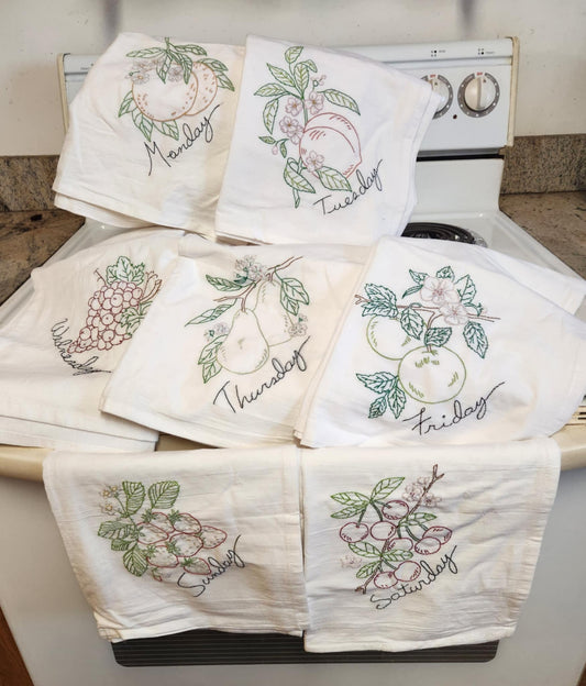 A set of white flour sack dish towels with different fruit designs and days of the week embroidered on them, displayed on a oven.