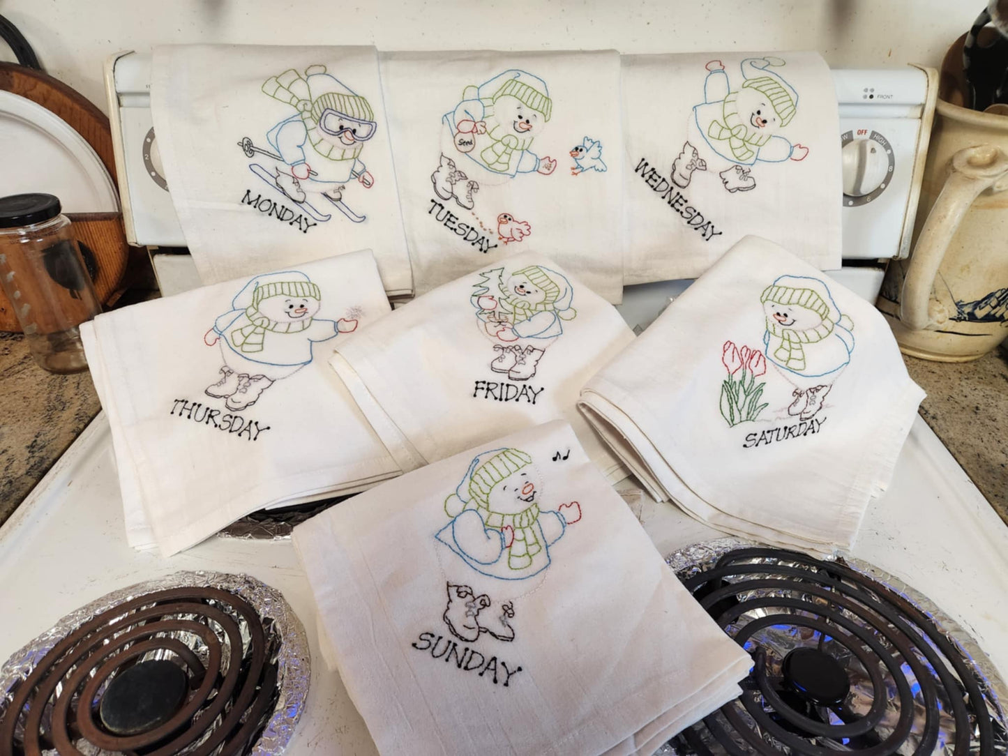 White dish towels with snowmen doing different activities embordered days of the week displayed on a oven