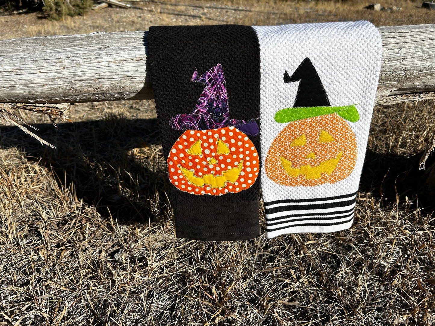 Two dish towels, one black the other white, has a orange jack o lantern but the pattern of fabric is different. They both are wearing Witches hats, ones purple and black the other is is black with a green rim  