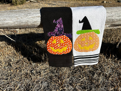 Two dish towels, one black the other white, has a orange jack o lantern but the pattern of fabric is different. They both are wearing Witches hats, ones purple and black the other is is black with a green rim  
