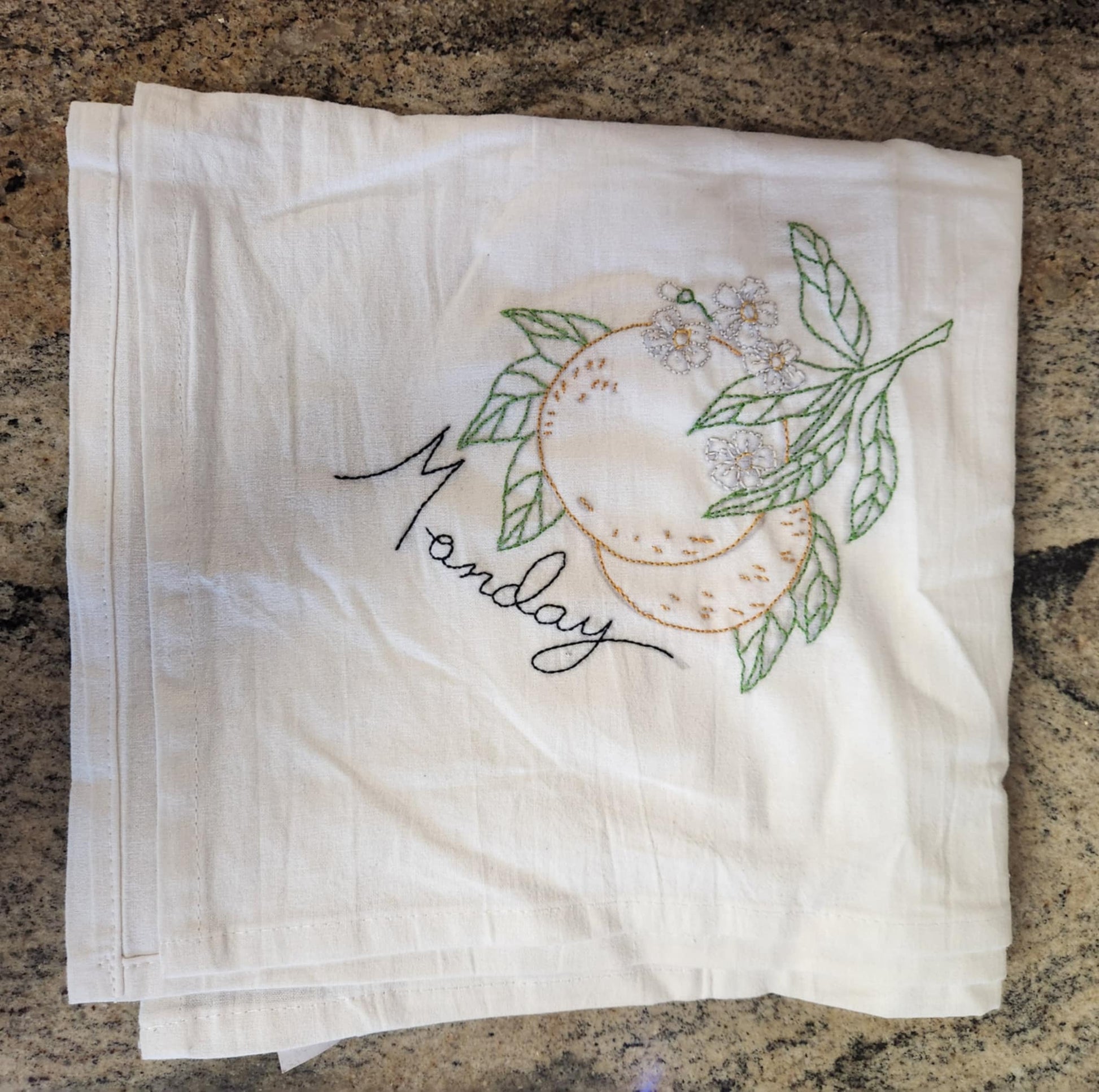 White dish towel with embroidery of a orange orange connected to a branch with green leaves and white flowers. "Monday" embroidered in black thread under the orange