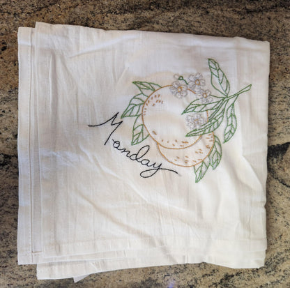 White dish towel with embroidery of a orange orange connected to a branch with green leaves and white flowers. "Monday" embroidered in black thread under the orange