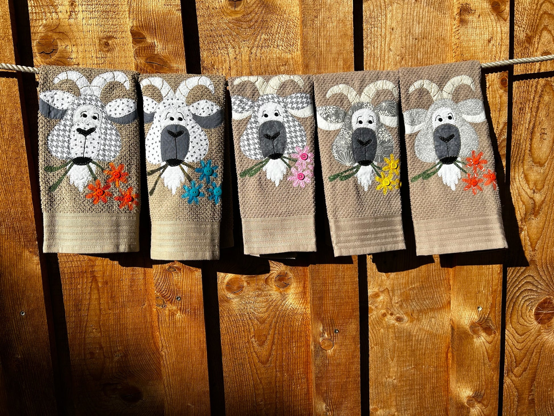 Five Tan hand towels with goats appliquéd on them each goat is made from different gray fabric. Each goat has 3 flowers in there mouth. Hanging on a clothes line