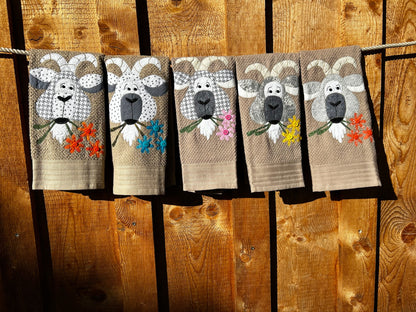 Five Tan hand towels with goats appliquéd on them each goat is made from different gray fabric. Each goat has 3 flowers in there mouth. Hanging on a clothes line