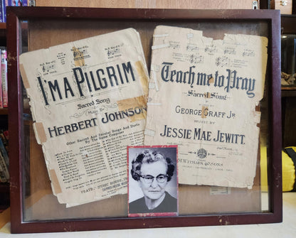 A wood shadow box with two sheet music ones I'm a pilgrim and the other Teach me to pray both look old and warn. There's a photo of a older woman in the center