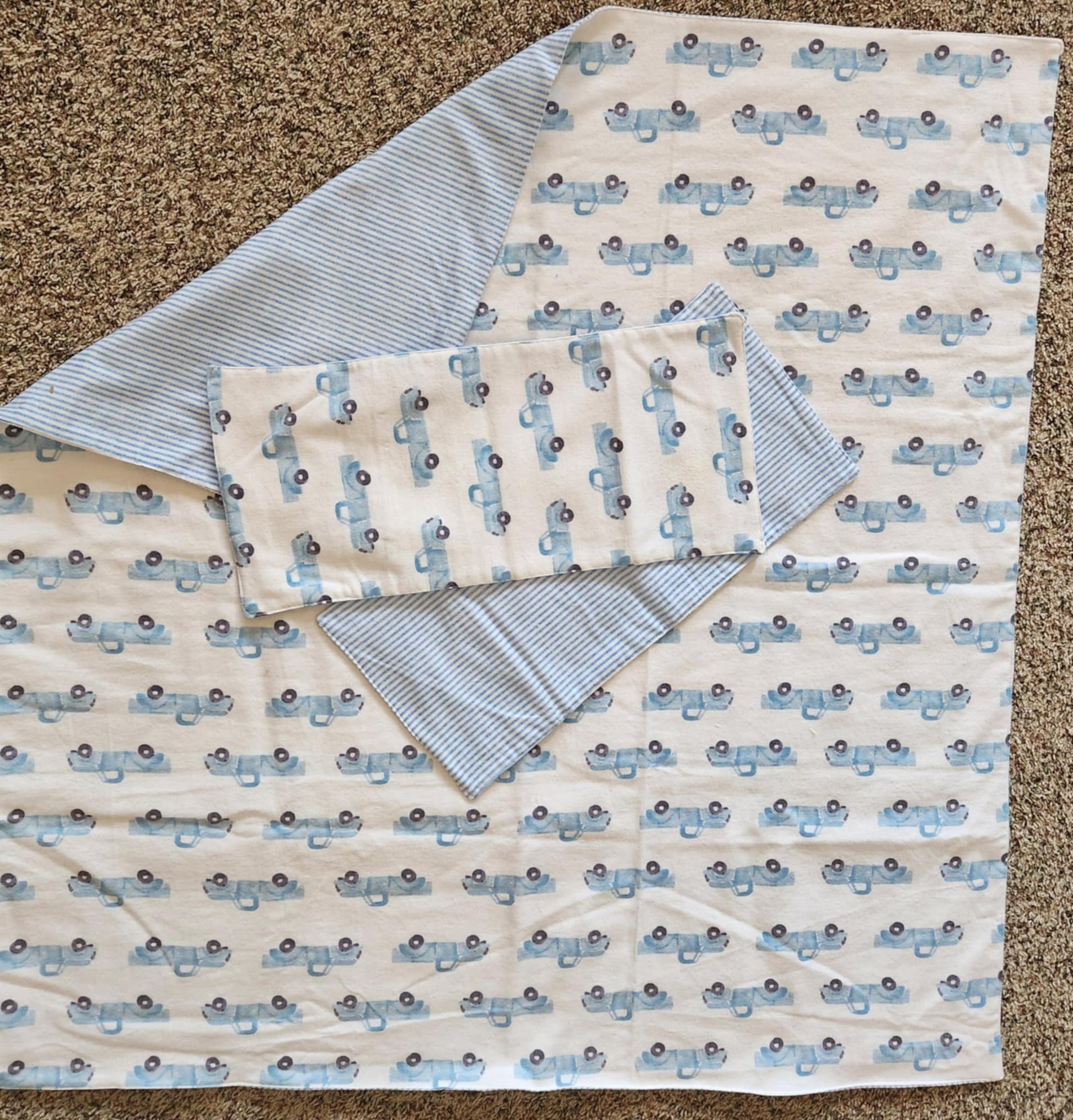 Square Receiving Blanket with Baby blue Pick up trucks with white background Fleece. With corner turned down showing baby blue and white Strip fleece. With tow burp rags  made from the same fabric on top of the blanket