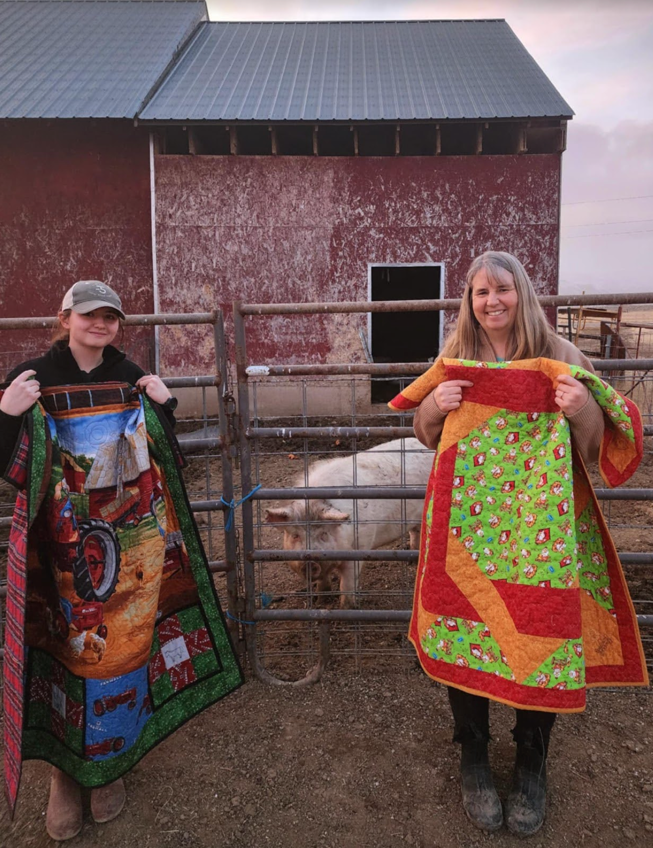 Two woman holding quilts with a pig in a pen between them