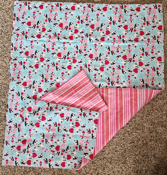 Square Receiving Blanket with Penguins, red and pink hearts with a blue background Fleece. With corner turned down showing pink, red, and white striped background fleece. With two burp rags made from the same fabric on top of the blanket 