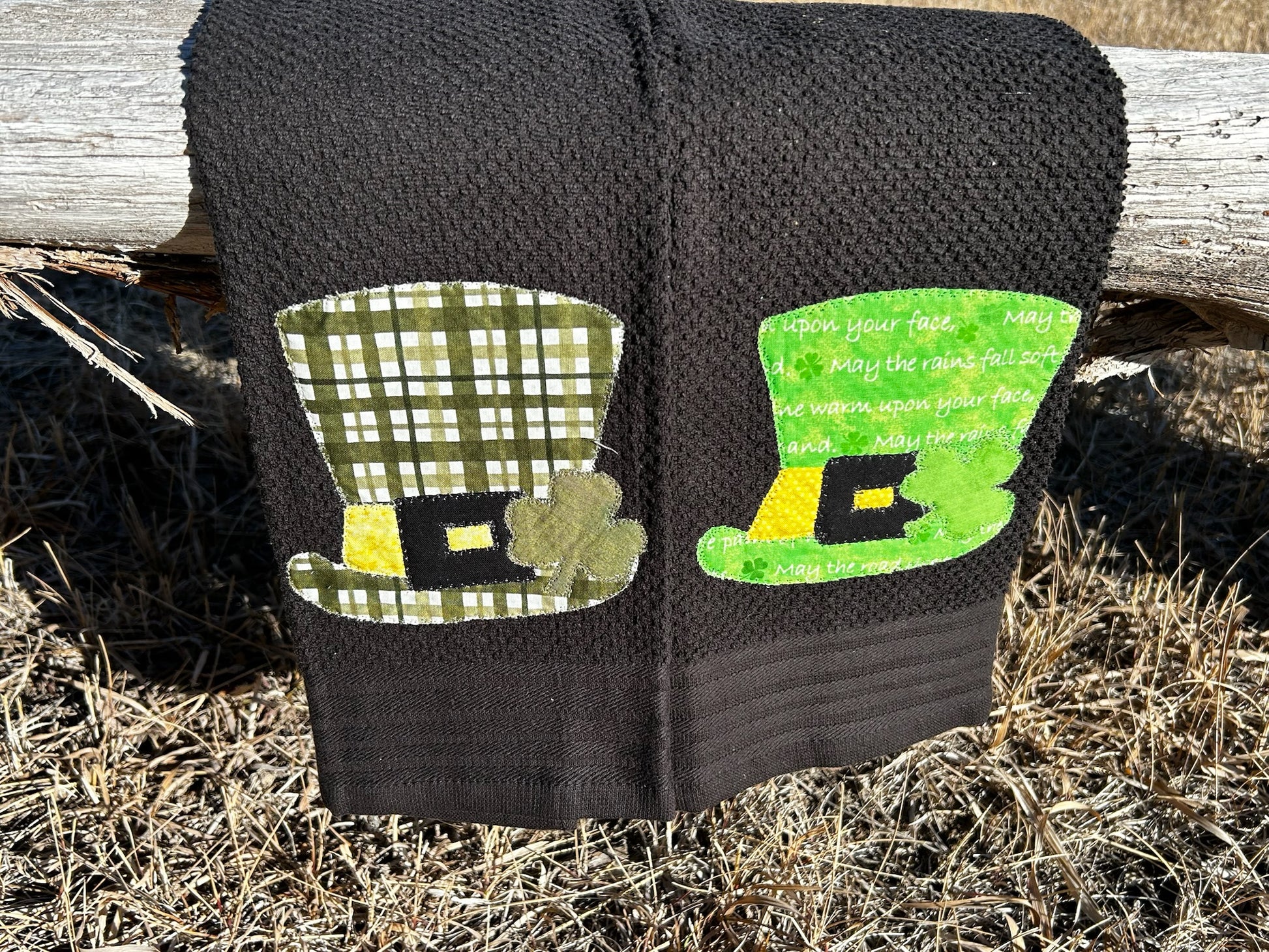 Two black hand towels has a leprechaun hat with a shamrock and buckle but each hat is a different shade of green 