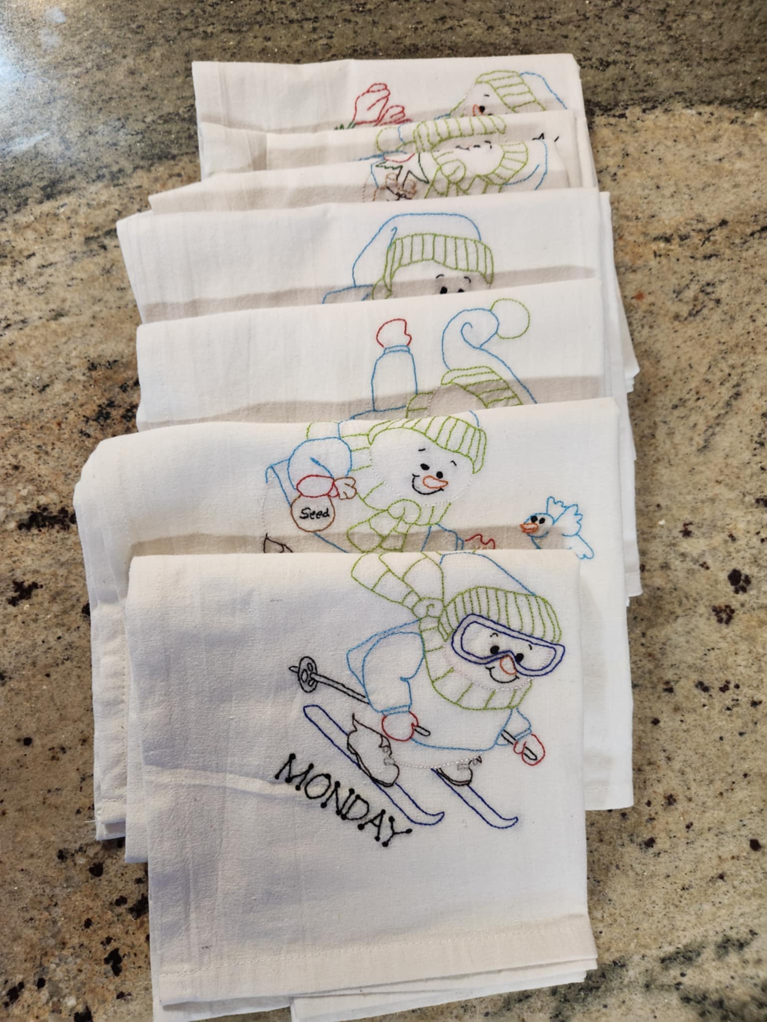 Seven white dish towels folded staggered towels with Snowman Embordered on them.  