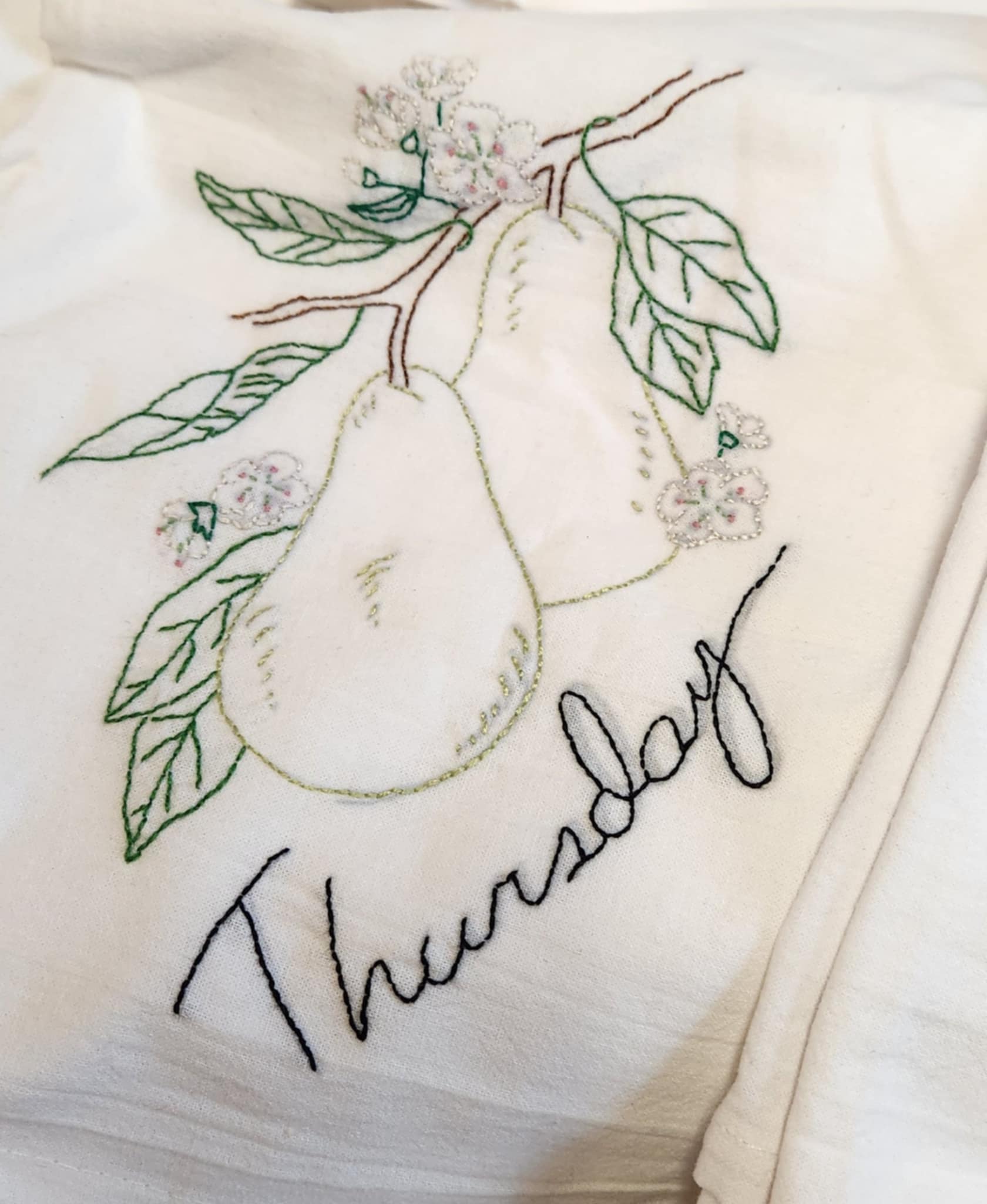 White dish  towel with embroidery of a green pear tree with to pears on a branch with green leaves and white flowers. "Thursday" Embroidered in black thread under the pears