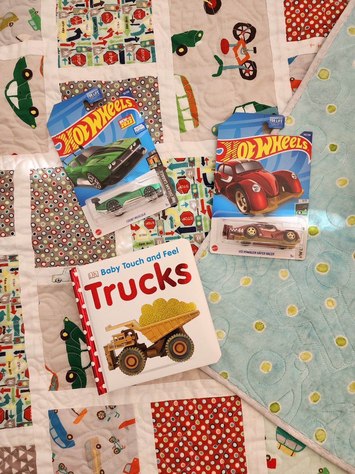 The quilt is primarily composed of rectangular patches with different car-themed fabrics. Some patches show repeating patterns of vehicles, others highlight individual cars or trucks, and some are simple geometric designs in red, gray, and beige. a corner folded back to reveal a light blue backing fabric with a subtle polka dot pattern. Two Hot Wheels car packages and a children's book titled "Trucks" are placed on top of the quilt.