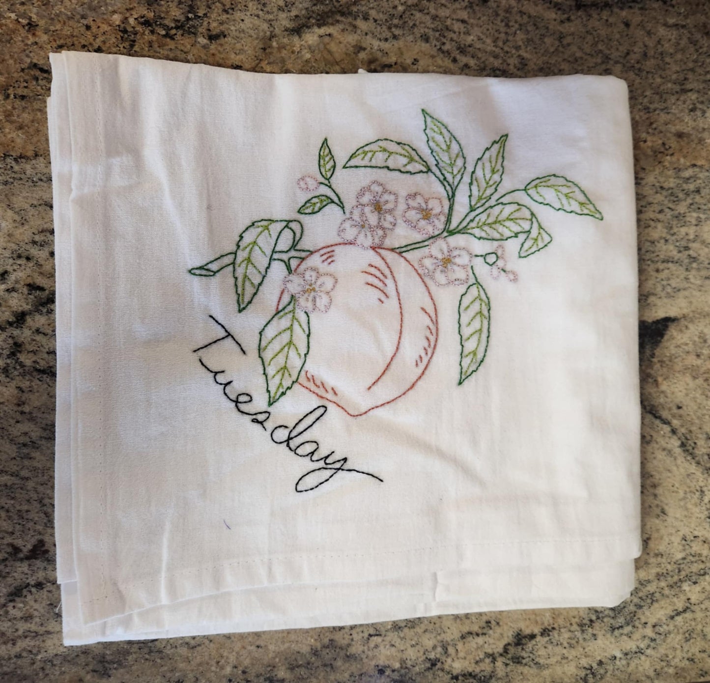 White dish towels with embroidery of a peach tree with a peach connected to a branch with green leaves and white flowers. "Tuesday" embroidered in black tread under the peach