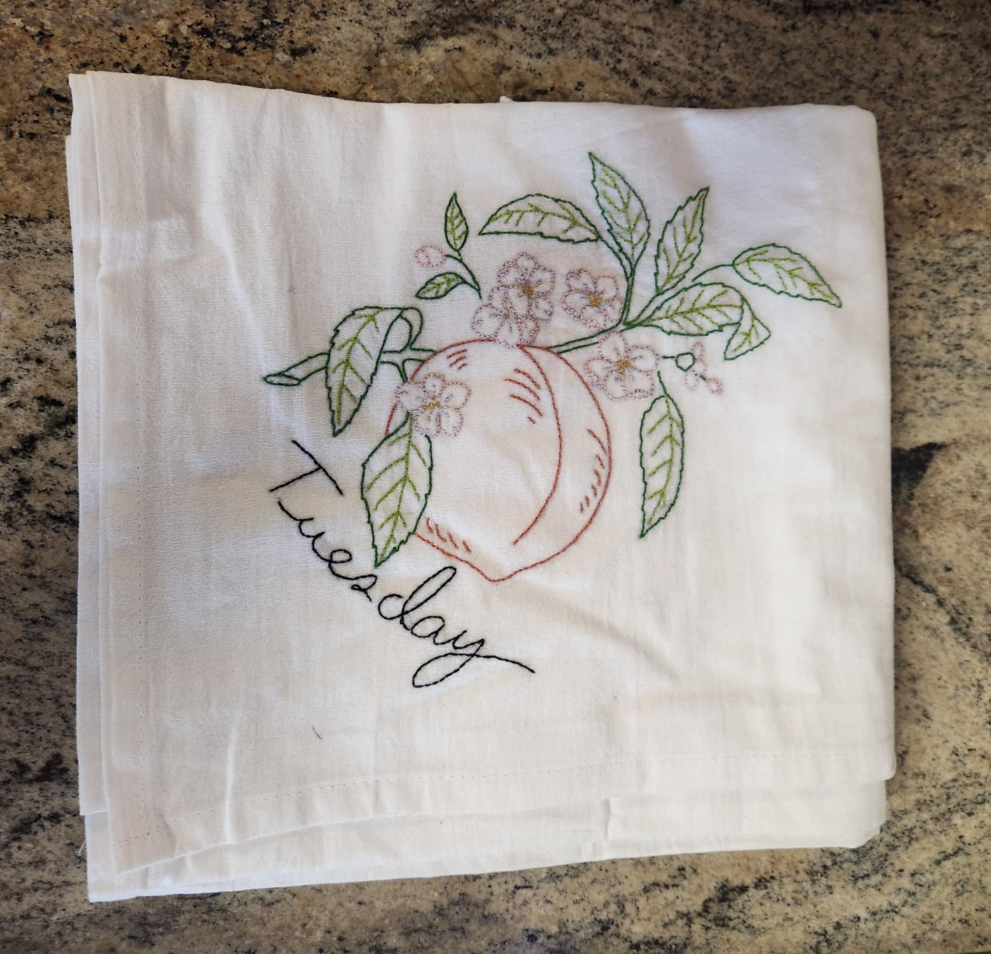 White dish towels with embroidery of a peach tree with a peach connected to a branch with green leaves and white flowers. "Tuesday" embroidered in black tread under the peach