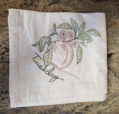White dish towels with embroidery of a peach tree with a peach connected to a branch with green leaves and white flowers. "Tuesday" embroidered in black tread under the peach