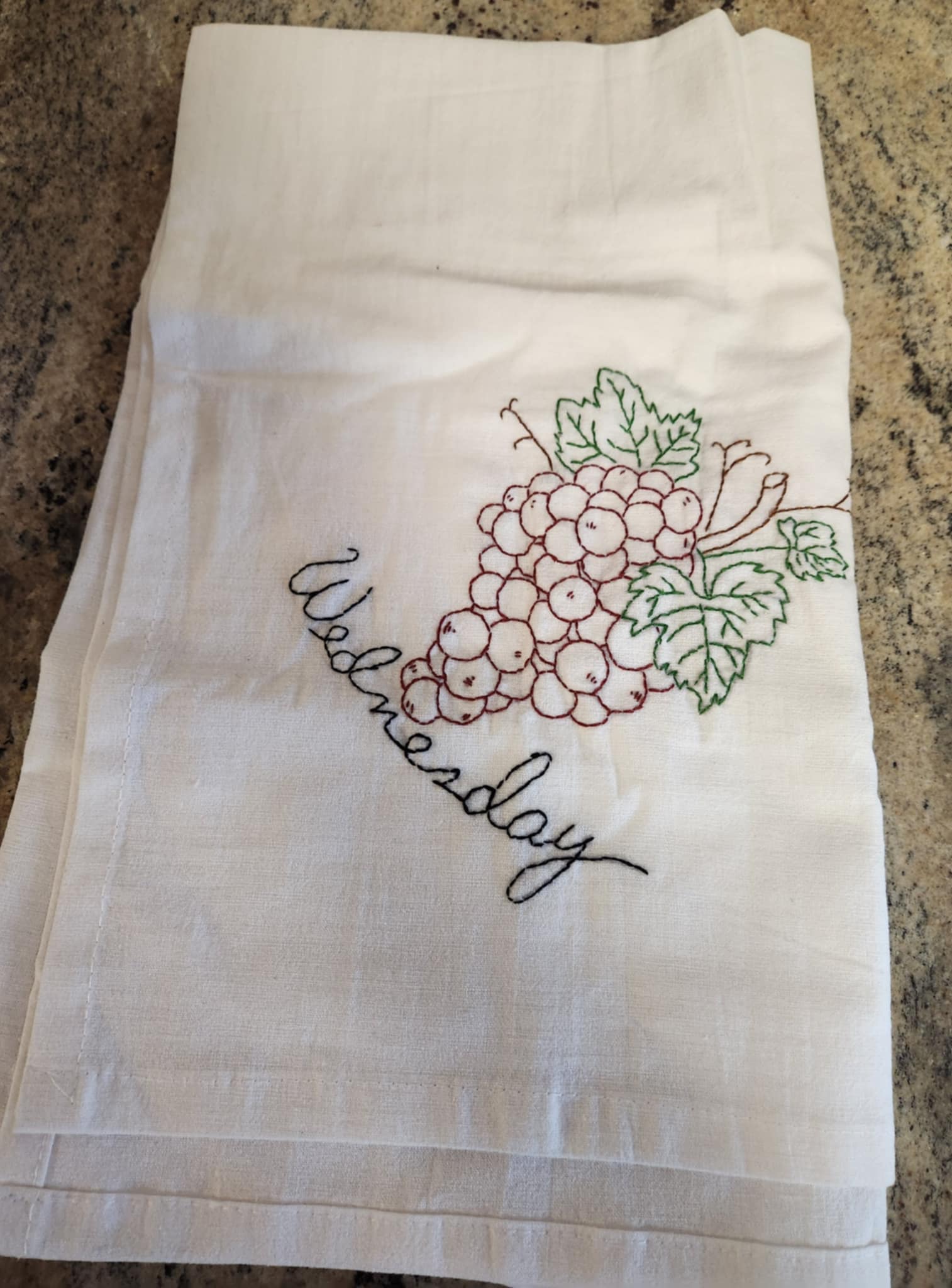 White dish  towel with embroidery of bunch of purple grapes on a vine with green leaves. "Wednesday" embroidered in black thread under the grapes 
