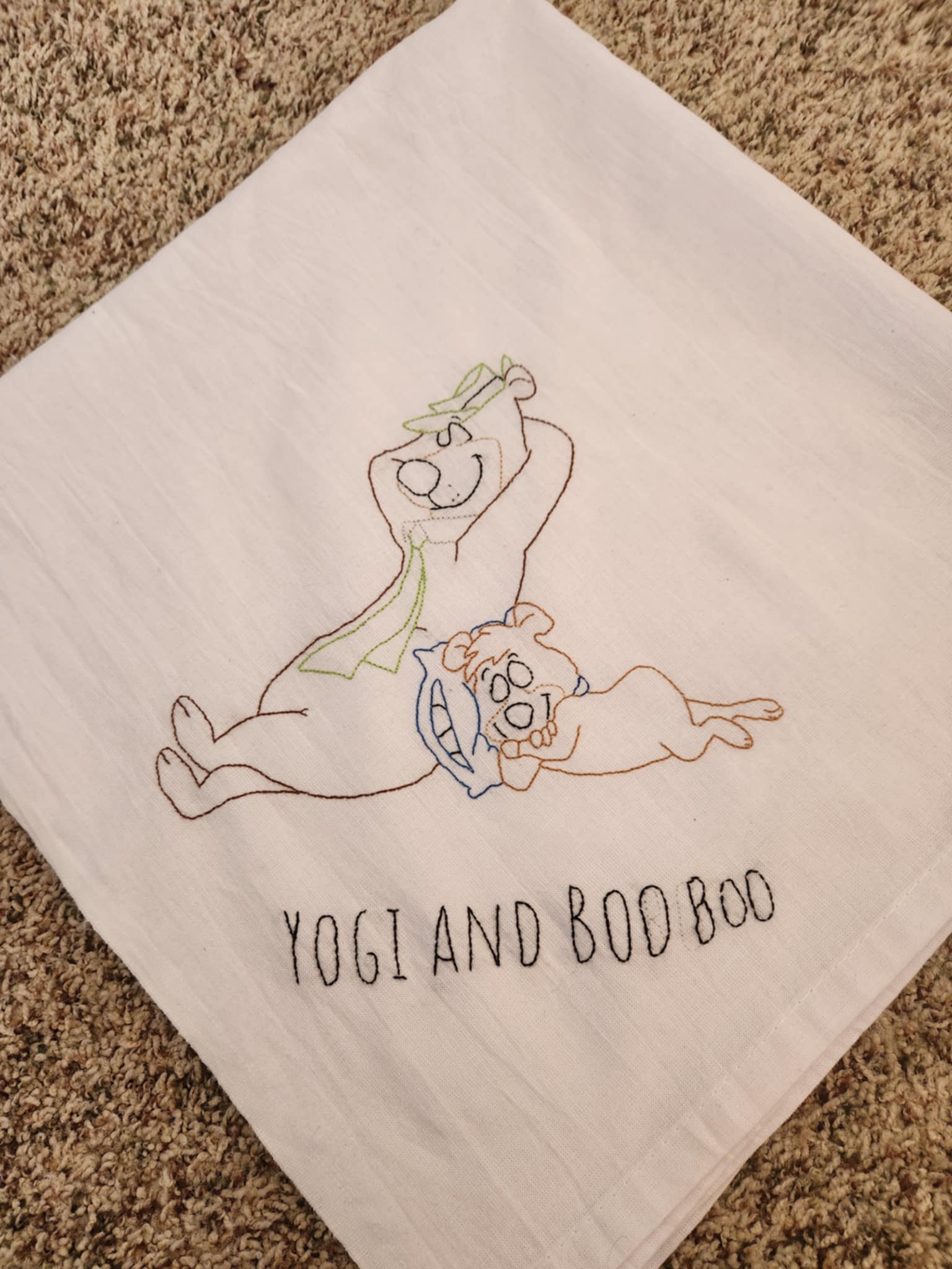 white towel folded in to a square with two bears sleeping, the small one resting on a blue pillow leaning on the bigger one. With the words "yogi and booboo" under them 